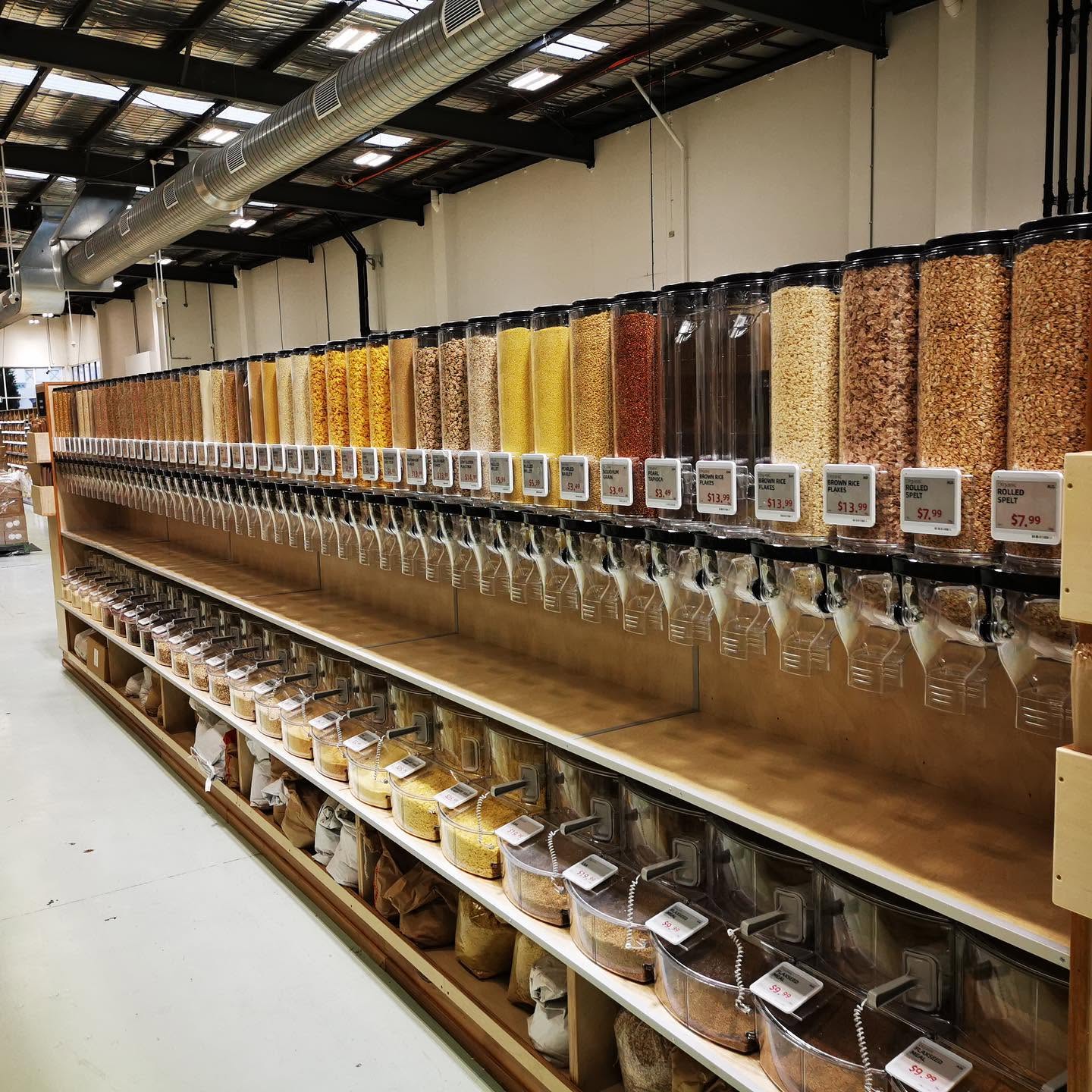 The bulk aisle at Terra Madre — a long wall of gravity dispensers filled with grains, flakes and cereals, price tags on every canister, with tubs of loose goods on the shelves below.