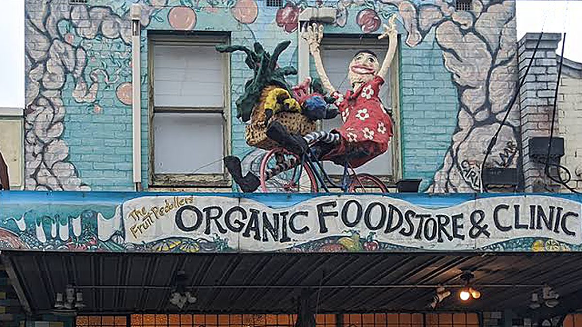 The original shopfront art in Northcote, up close — two sculpted figures riding a bicycle on the painted facade, above the hand-painted sign reading The Fruit Peddlers Organic Foodstore & Clinic.