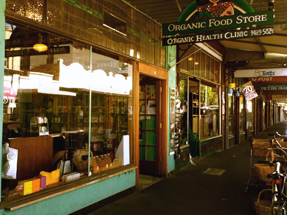 The High St shop under its verandah — a hanging sign reading ORGANIC FOOD STORE over ORGANIC HEALTH CLINIC, leadlight windows and a green tiled stallriser.