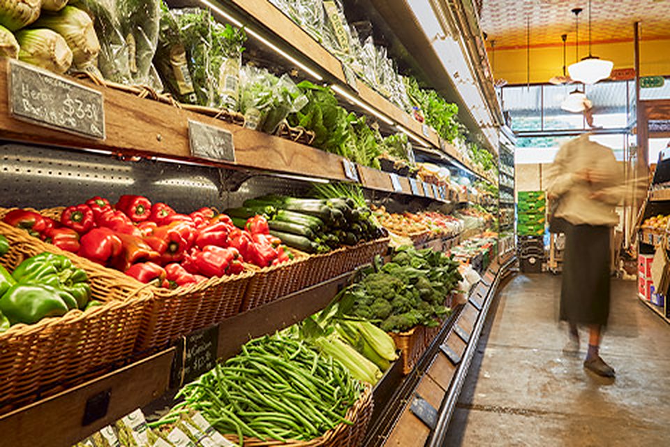 The produce aisle at Terra Madre — wicker crates of red and green capsicums, zucchini, broccoli, beans and celery under a timber shelf of leafy greens and herbs, with a chalked price card, and a customer walking past at the far end.
